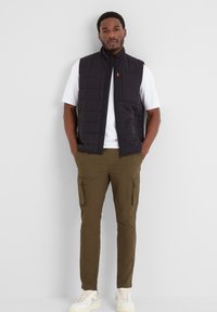Black quilted vest with a zippered front and chest pocket, worn over a white t-shirt and olive cargo pants, paired with off-white sneakers.