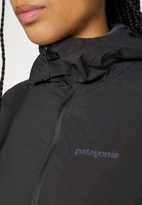 Close-up de uma pessoa a usar uma jaqueta preta da Patagonia com um colarinho alto e um fecho visível à frente.