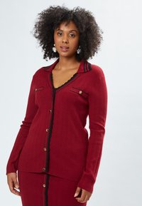 Roter Cardigan mit geripptem Textur, schwarzen Akzenten, einem geschwungenen Kragen und goldfarbenen Knopfdetails entlang der Vorderseite und Taschen.