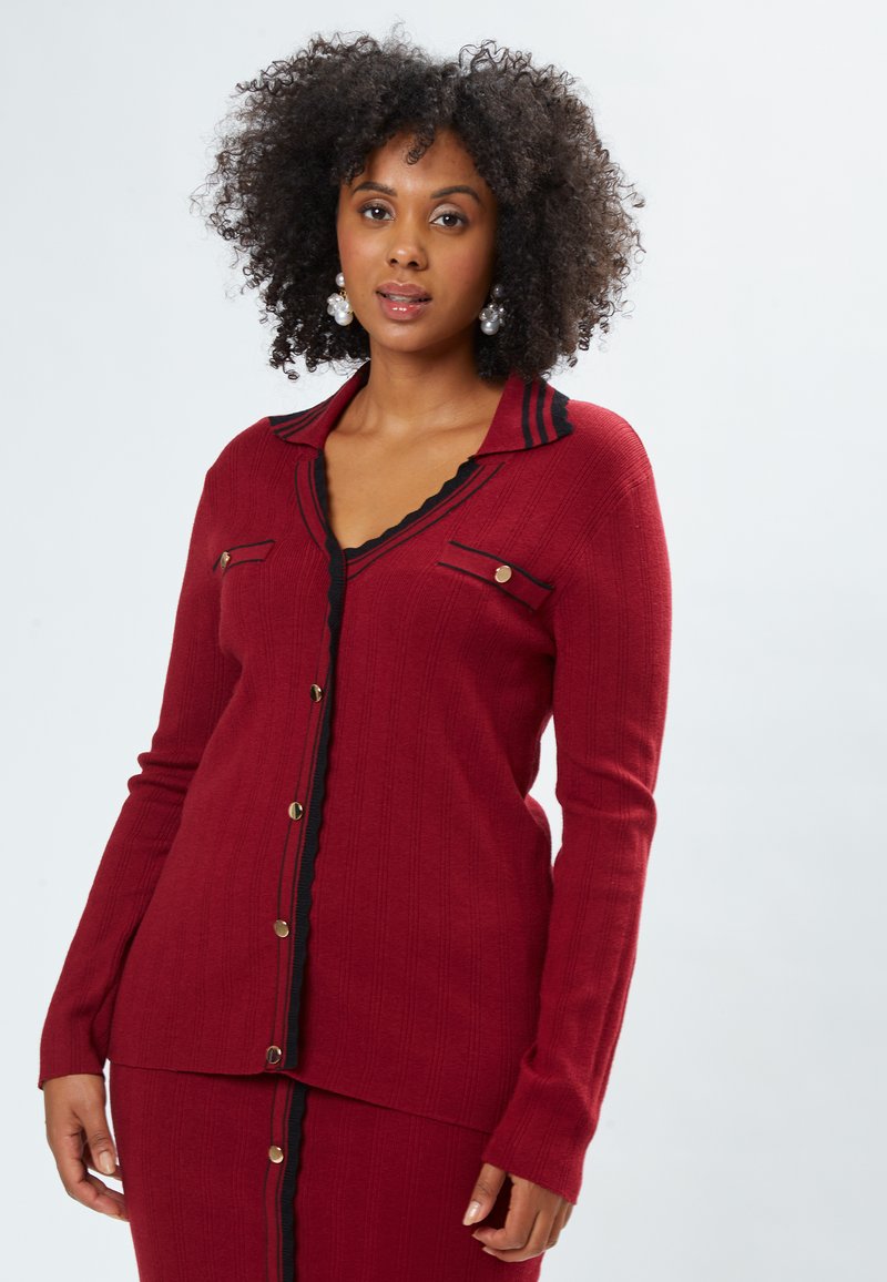 Roter Cardigan mit geripptem Textur, schwarzen Akzenten, einem geschwungenen Kragen und goldfarbenen Knopfdetails entlang der Vorderseite und Taschen.