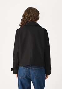 Černý krátký kabátek z vlněné směsi se širokým límcem, manžetami na knoflíky a hladkou texturou, kombinovaný s modrými džínami.