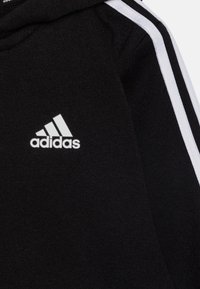 Felpa nera con cappuccio, caratterizzata da strisce bianche sui lati e un logo Adidas bianco sul davanti, realizzata in un morbido tessuto testurizzato.
