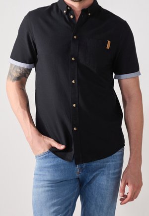 Camisa negra de manga corta con cuello abotonado, puños azules enrollados, un bolsillo en el pecho y botones en tono dorado. Presenta una textura de tela suave.