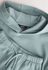 Hele sinine tekstuuriga kapuutsiga dressipluus ja sobivad dressipüksid. Omavad kapuutsi, paelaga reguleeritavat vööd ning pehmet vahvlilõikelist kangast. Brändi silt on näha.