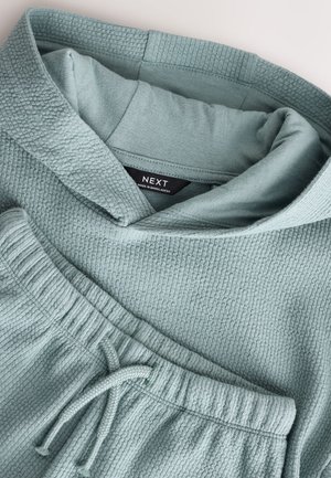 Sweat à capuche bleu clair texturé et joggers assortis. Comprend une capuche, une taille à cordon de serrage, et un tissu doux en maille gaufrée. Étiquette de marque visible.