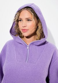 Žena s vlnitými blond vlasy, oblečená v levandulové fleecové mikině s polovičním zipem, hledící mírně na stranu na bílém pozadí.