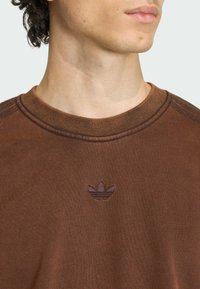 Hnedá bavlnená mikina s rebrovaným výstrihom a malým vyšívaným logom na hrudi. Hladká textúra s kontrastnými švíkmi.