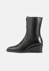 Črne usnjene gležnjarje z klinastim petam, ki imajo koničast nos in vezalni sistem ob strani, gladko teksturo z rahlim sijajem.