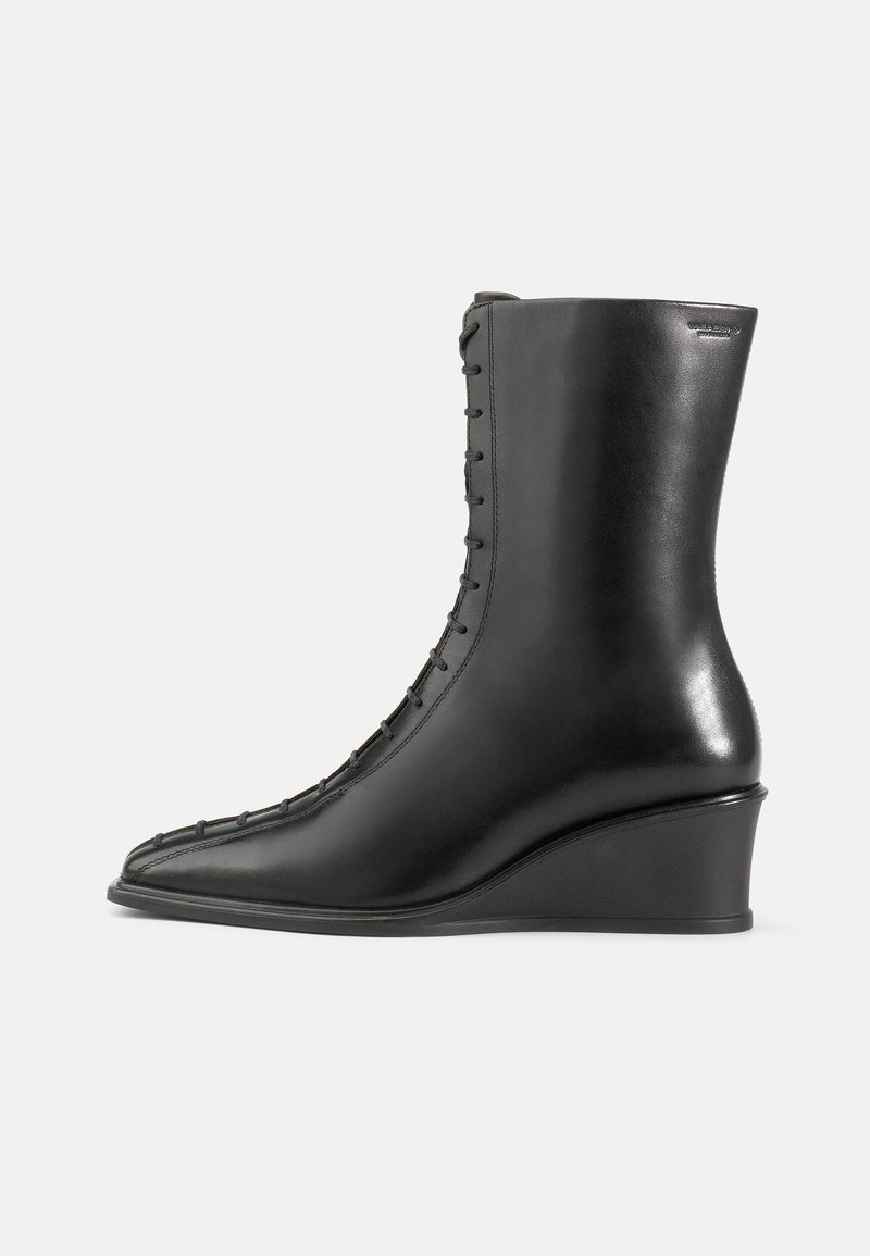 Črne usnjene gležnjarje z klinastim petam, ki imajo koničast nos in vezalni sistem ob strani, gladko teksturo z rahlim sijajem.