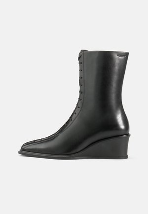 Black leather ankle boot with a wedge heel, featuring a pointed toe and a side lace-up design with stitched detailing. Smooth texture.