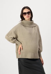 Pull beige surdimensionné avec un col haut à côtes et des manches larges. Tissu en tricot texturé ; associé à une jupe noire brillante.