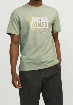 Man draagt een lichtgroen Jack & Jones T-shirt met kaartontwerp en donkergrijze broek, staat met één hand in de zak.