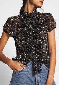 Blouse noire transparente avec un devant à volants, motif à pois en beige clair, manches courtes et fermetures à boutons, associée à un jean en denim clair.