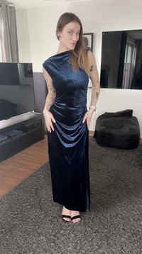 Vestido de veludo azul escuro com decote assimétrico, detalhes franzidos e uma silhueta longa e fluida. Usado com sandálias pretas de tiras.