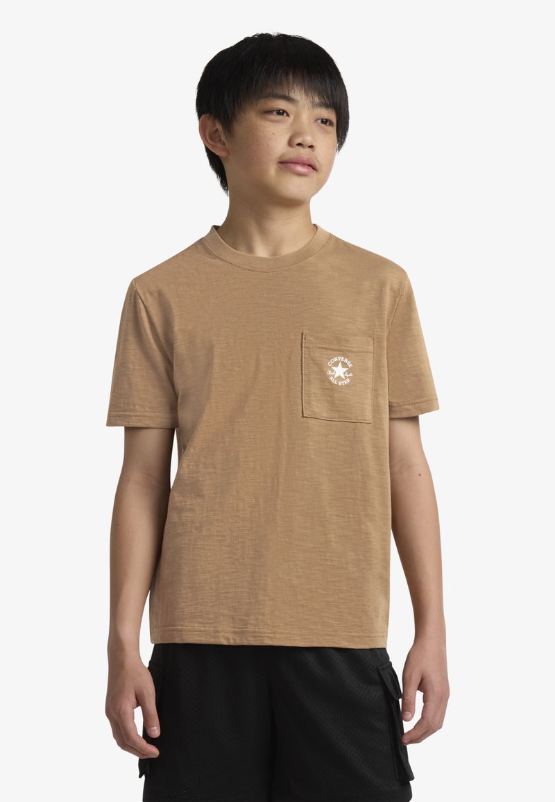 Tan kratka majica z rokavi, z teksturirano zaključkom, na levi strani prsnega žepa in belim tiskanime logotipom na žepu. Nosi se s črnimi kratkimi hlačami.