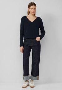Granatowy sweter z prążkowanego dzianiny z dekoltem w serek, zestawiony z ciemnoniebieskimi, szerokimi dżinsami z podwiniętymi mankietami oraz białymi tenisówkami z brązowymi akcentami.