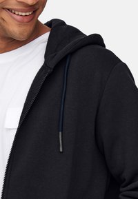 Schwarze Zip-Hoodie mit weicher Textur, ausgestattet mit einer Kapuze mit Kordelzug, kontrastierenden marineblauen Schnürsenkeln und einem weißen T-Shirt darunter.