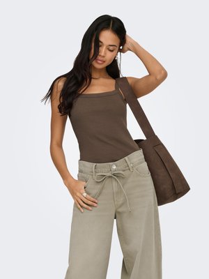 Joven mujer con cabello largo y oscuro que lleva una camiseta sin mangas marrón y pantalones beige de pierna ancha, llevando un gran bolso marrón al hombro.