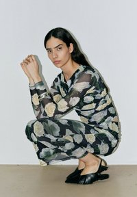 Zwart doorzichtig outfit met witte en groene rozen, featuring lange mouwen en een getailleerd silhouet. Zwarte franjesandalen.