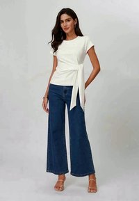 Bílý krátký rukáv top s bočním zavázáním, kombinovaný s širokými modrými džíny. Outfit má uvolněný střih a hladké textury.