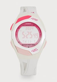 Montre Casio numérique avec un boîtier blanc, des accents roses et un bracelet texturé. Présente un grand affichage indiquant l'heure et la date. Autonomie de batterie de dix ans.