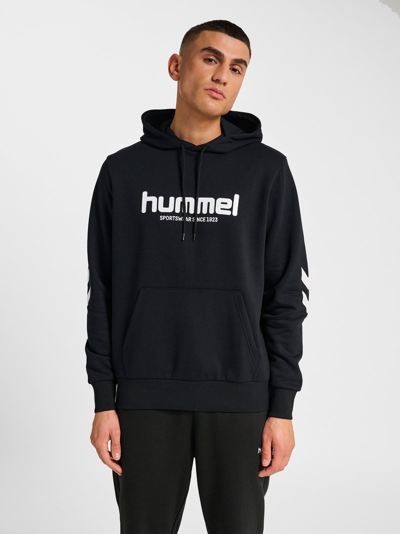Man draagt een zwarte hummel hoodie met wit logo en zwarte broek, staand tegen een effen lichtgrijze achtergrond.