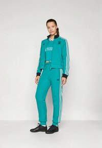 Sportssdress i blågrønn med hvite striper, kort jakke og lange bukser. Har rund hals, ribbede mansjetter og logo-detaljer.