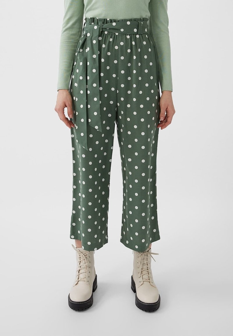 Pantalon vert taille haute à pois blancs avec une ceinture assortie, porté avec des bottines beiges à lacets et un haut vert clair à manches longues.