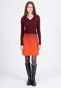 Femme debout, portant un haut à manches longues côtelé bordeaux, une jupe à motifs orange, des collants noirs et des bottes noires jusqu'aux genoux sur un fond blanc.