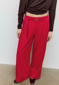 Pantaloni a gamba larga in tessuto rosso vivace, con vita incinturata e texture morbida. Abbinati a scarpe scure con tacco.