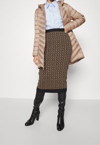 Polstret beige frakke, lyseblå skjorte, mønstret sort pencilnederdel med guldaccents og sorte knæhøje støvler med hæl, stående på grå baggrund.