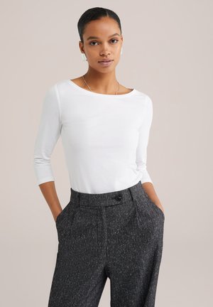 Femme les mains dans les poches portant une chemise blanche à manches longues, un pantalon gris texturé taille haute, un collier en argent et des boucles d'oreilles blanches, fond neutre.