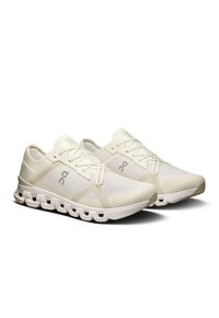 Baskets de sport beige clair avec tige en mesh, lacets plats et accents de logo. La semelle rembourrée possède des découpes circulaires pour la flexibilité et le soutien.