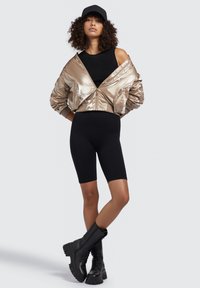 Metalická zlatá bomber bunda, černý žebrovaný top, těsné černé šortky a černé vysoké boty po kolena, s černou kšiltovkou, na neutrálním pozadí.