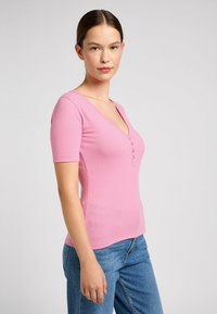 Lyserød ribbet kortærmet top med V-hals og fem-knaps placket; tætsiddende design fremhæver formen; glat tekstur. Parret med denim jeans.