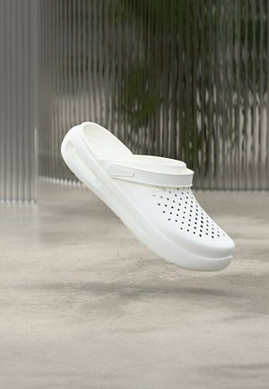 Sabot blanc ventilé à enfiler avec bride arrière flottant au-dessus d'un sol en béton lisse et un fond de stores verticaux texturés.