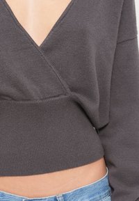 Γκρι cropped πουλόβερ με βαθύ V-λαιμόκοψη, με υφή πλεκτή και μακριά μανίκια, σε συνδυασμό με ανοιχτό μπλε τζιν.
