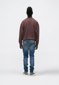 Mężczyzna z dredami, ubrany w brązową bluzę, niebieskie dżinsy i szare trampki, stoi tyłem na jednolitym, białym tle.