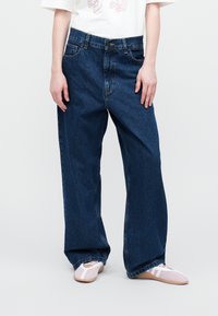 Jeans de mezclilla azul de pierna ancha con cintura alta, que cuentan con bolsillos y un diseño clásico de cinco bolsillos. Usados con zapatos de lona rosas.