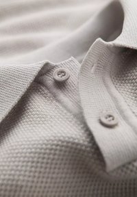 Světle šedé polo tričko s texturovanou tkaninou, dvěma knoflíky na límci a tvarovaným výstřihem. Hladký povrch a minimální design.