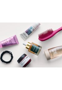 En platt bild av skönhetsprodukter: en rosa utredningsborste, olika krämrör i nyanser av lila och vitt, en oljeflaska med guldaccent, och ett svart hårband.