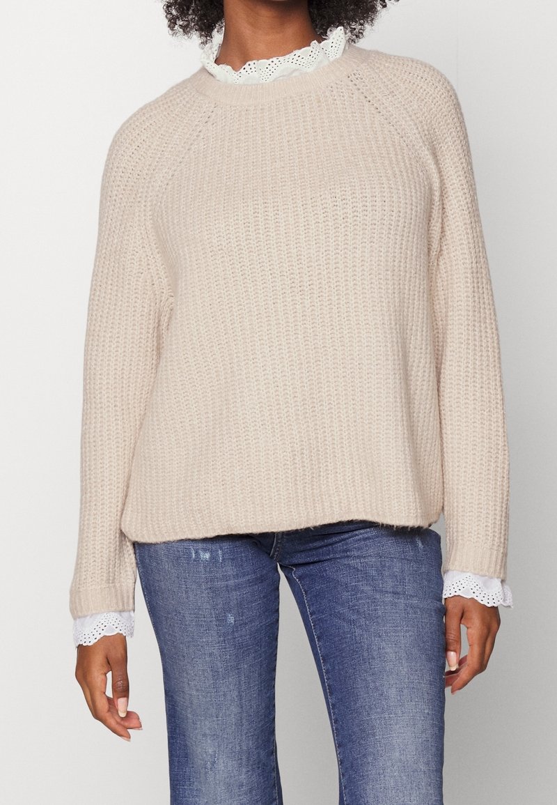 Femme portant un pull en maille gaufrée beige superposé sur un chemisier blanc orné de dentelle œillet et un jean bleu délavé, debout devant un fond uni.