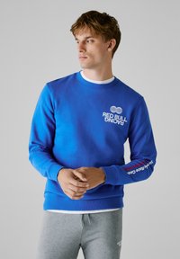 Νέος άνδρας με μπλε φούτερ Red Bull Racing και γκρι φόρμες, με τα χέρια ενωμένα, κοιτάζοντας στο πλάι μπροστά σε έναν απλό γκρι φόντο.