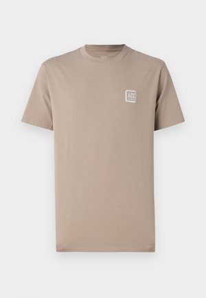 Camisa de algodón beige con cuello redondo, mangas cortas y un pequeño parche de logo blanco en el lado izquierdo del pecho. Textura suave, ajuste casual.