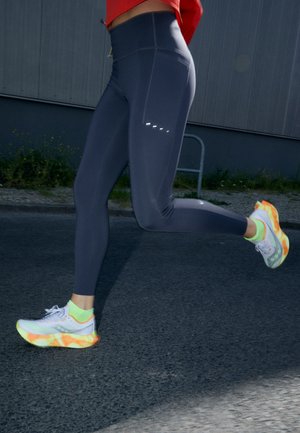 Donkerblauwe leggings met een hoge tailleband, voorzien van reflecterende details. Gekoord met witte en neonoranje hardloopschoenen.