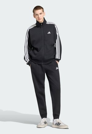 Joven con las manos en los bolsillos, llevando un chándal negro de Adidas con franjas blancas y zapatillas a juego sobre un fondo claro.