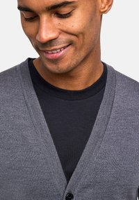 Grauer Cardigan mit V-Ausschnitt, gerippter Textur und einem einzelnen schwarzen Knopf, über einem schwarzen T-Shirt getragen. Nahaufnahme, auf Details fokussiert.