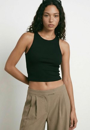 Jeune femme aux cheveux bouclés portant un crop top noir sans manches et un pantalon beige plissé, debout devant un fond clair uni.