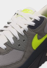 Gros plan d'une basket grise Nike avec des lacets blancs, un logo Swoosh jaune néon et des détails en mesh sur le côté et la languette.
