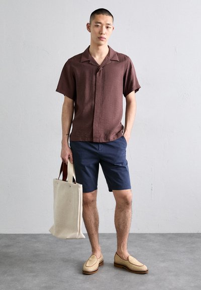 Chemise marron à manches courtes, shorts navy, slip-ons tissés beiges, et tote bag léger beige avec des poignées marron ; design et textures minimalistes.